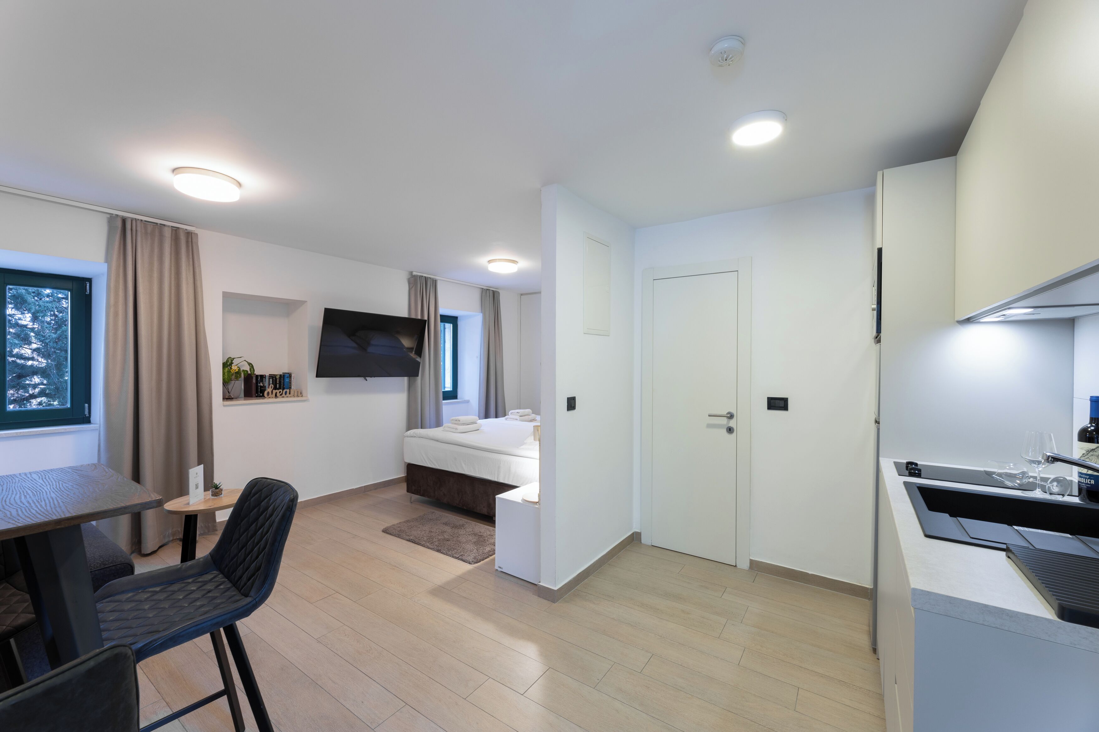 Una habitación de hotel moderna con una cama, un escritorio con silla, un televisor en la pared y una kitchenette.