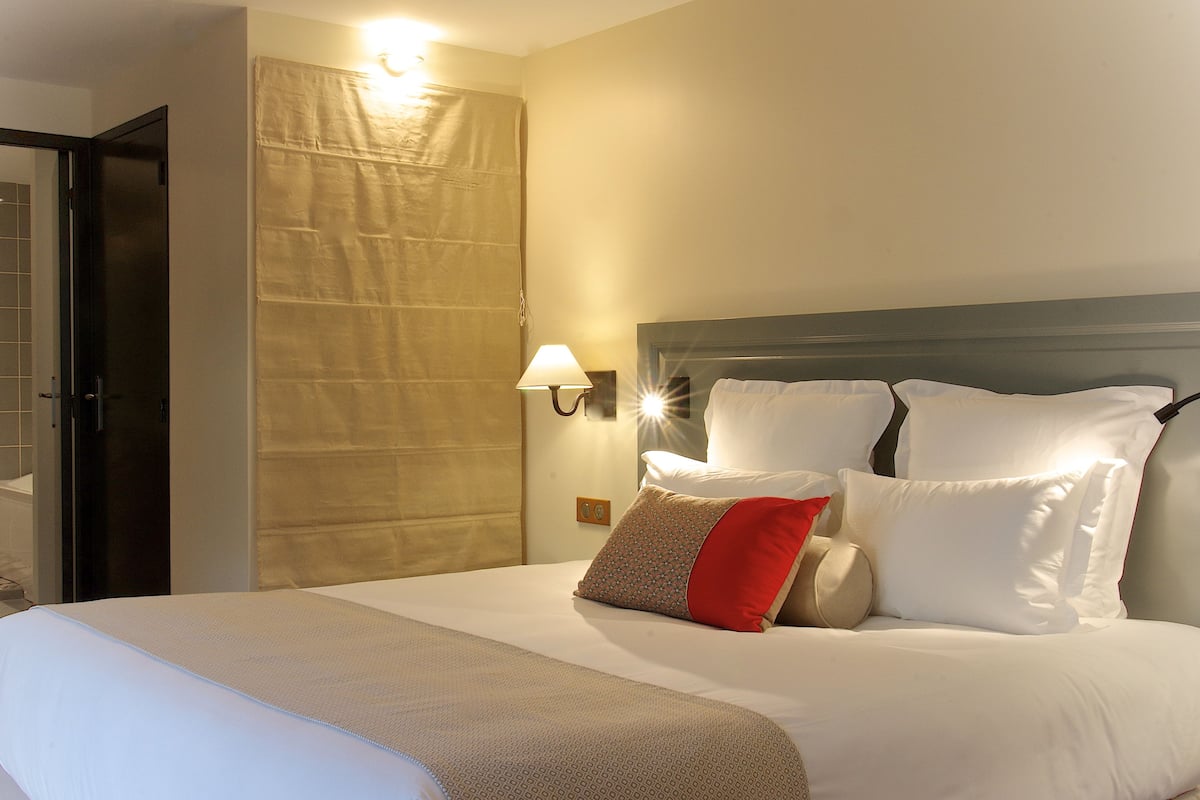 Una camera d'albergo con un letto grande, una testiera imbottita con numerosi cuscini, una lampada a parete e una tenda alla finestra.