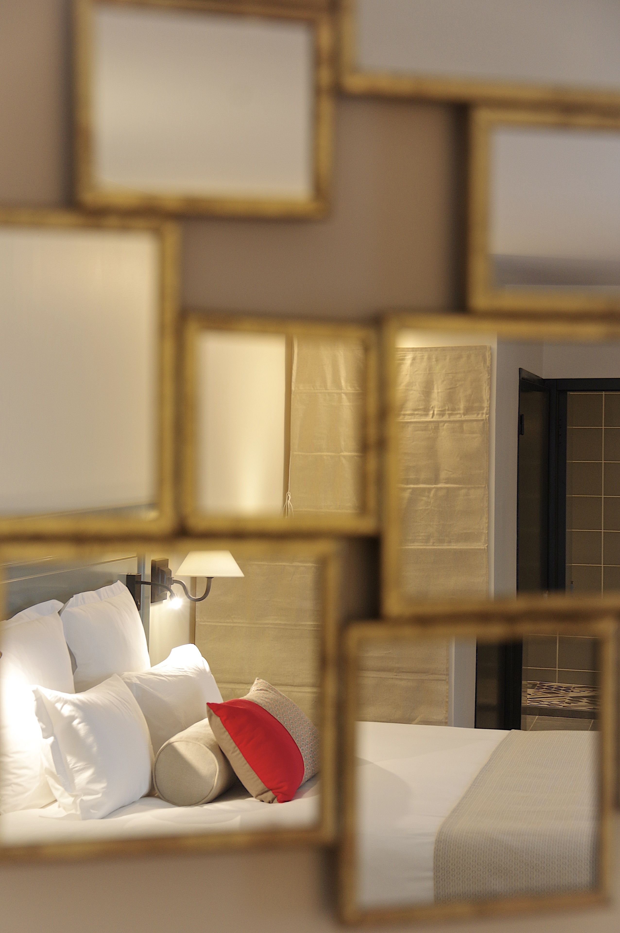 A hotel room with a bed, pillows, a lamp, and a mirror reflecting the room's interior.