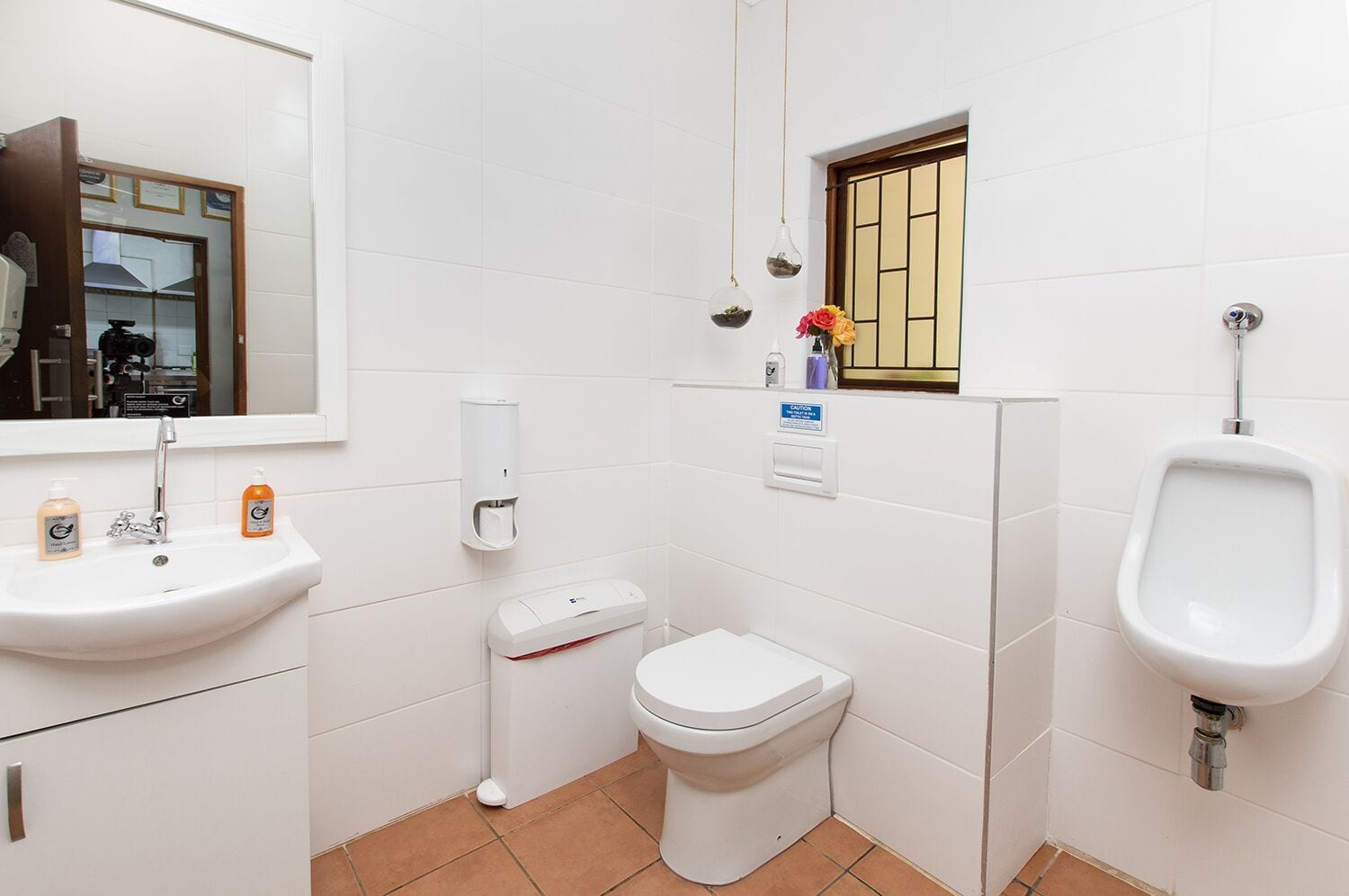 A small, clean bathroom with a sink, toilet, urinal, and a small window with a view.
