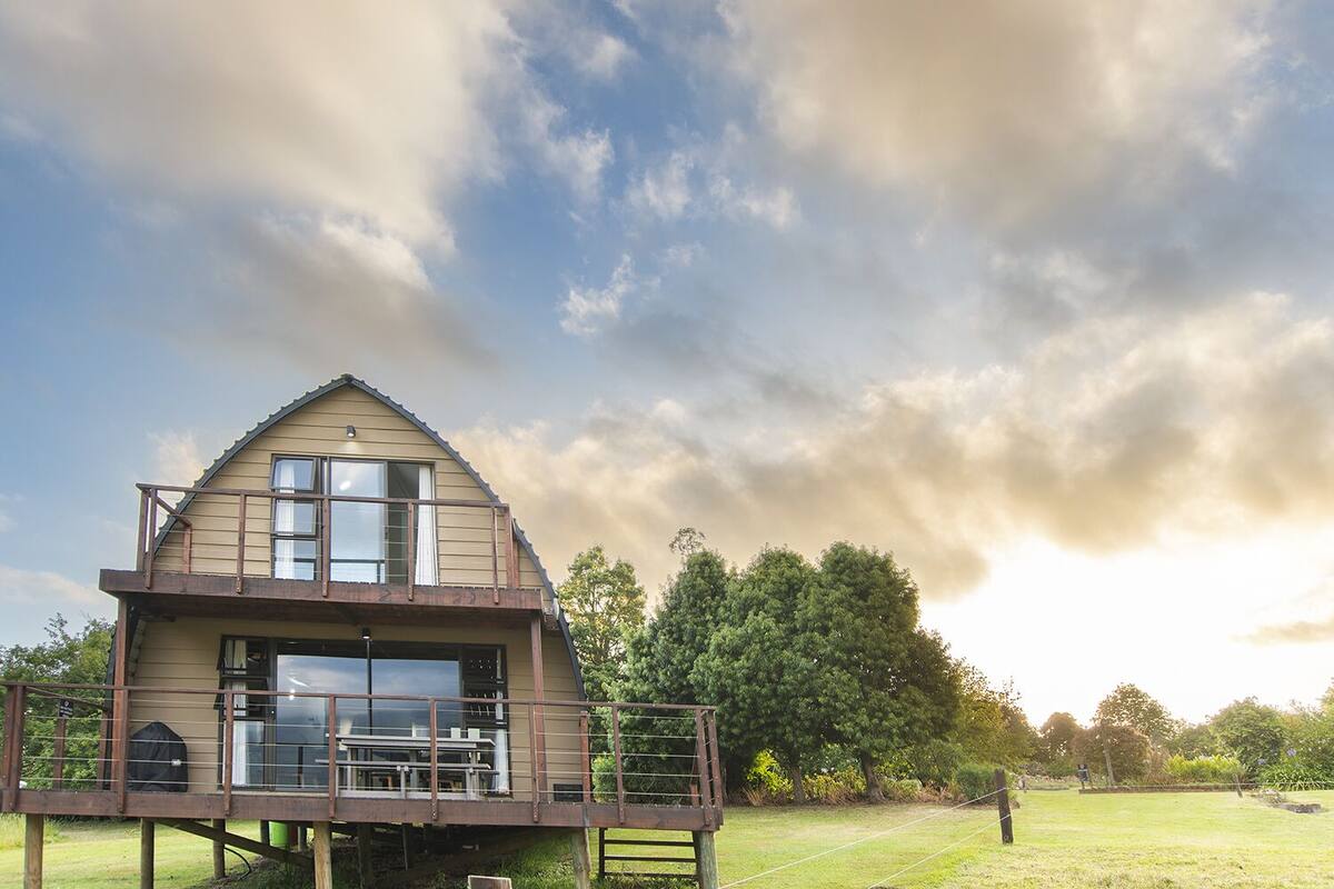 A two-story house with a balcony, a covered outdoor seating area, and a view of a grassy field and trees.