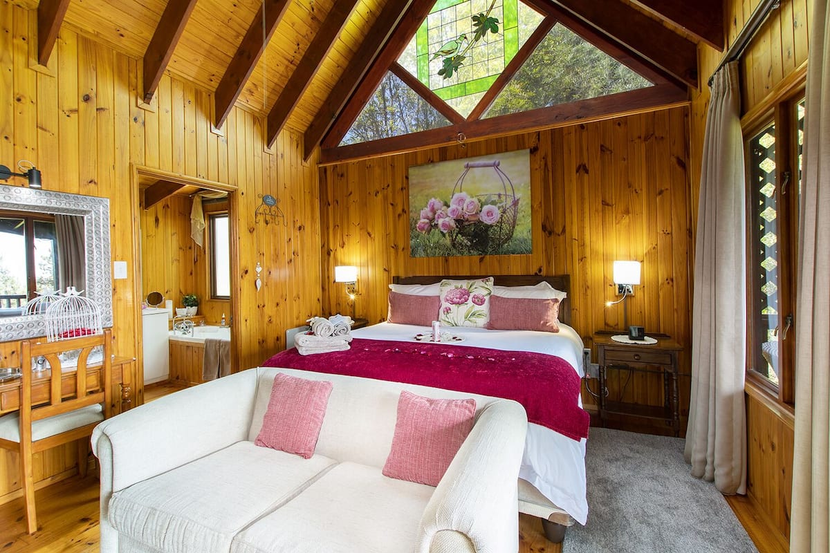 A wooden cabin bedroom with a bed, a chair, a desk, and a painting on the wall.