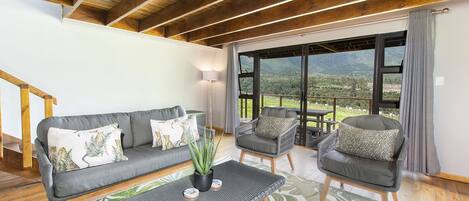 A modern living room with a grey sofa, two armchairs, a coffee table, and a view of a mountainous landscape through large windows.