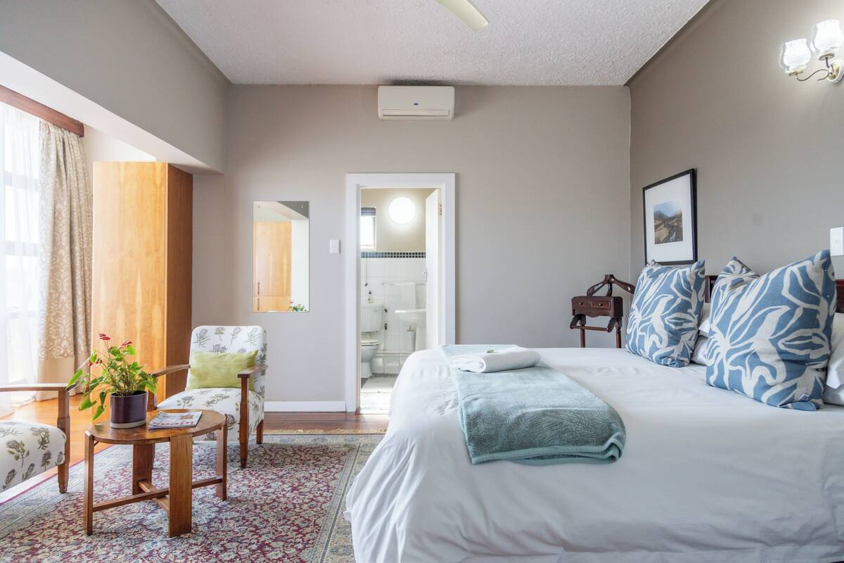 Ein Schlafzimmer mit Bett, Stuhl, kleinem Tisch mit Pflanze, Spiegel und einem durchgegangenen Tür angedeuteten Badezimmer.