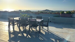 A rooftop terrace with a table and chairs, potted plants, and a view of the ocean and hills.