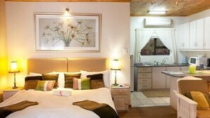 A bedroom with a large bed, bedside lamps, a framed floral artwork, and a view of a kitchen with a window. - Cul de Sac (Oudtshoorn)