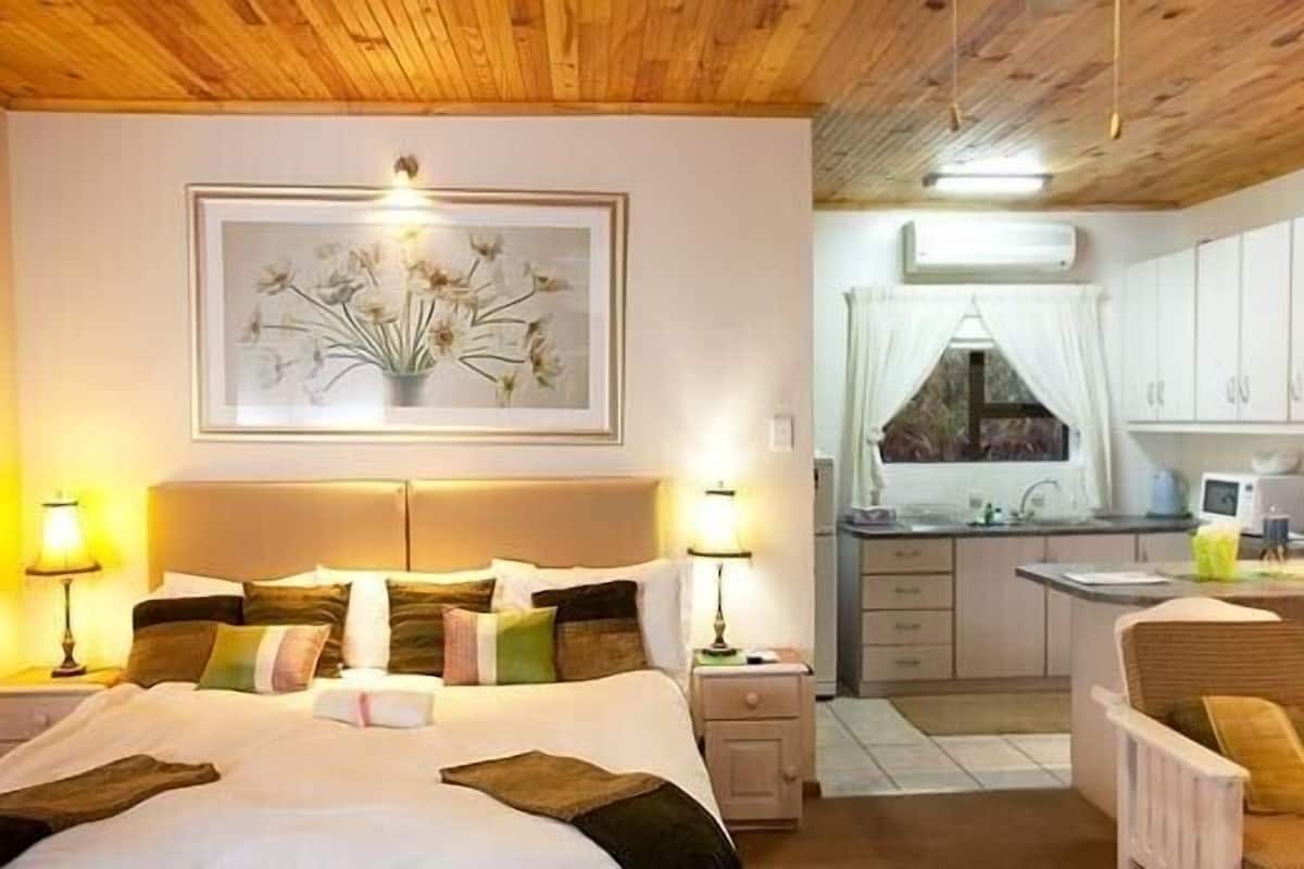 A bedroom with a large bed, bedside lamps, a framed floral artwork, and a view of a kitchen with a window.