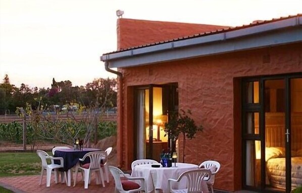 A building with a red exterior, a patio with white plastic chairs and a table, and a large sliding glass door leading to a room with a bed.