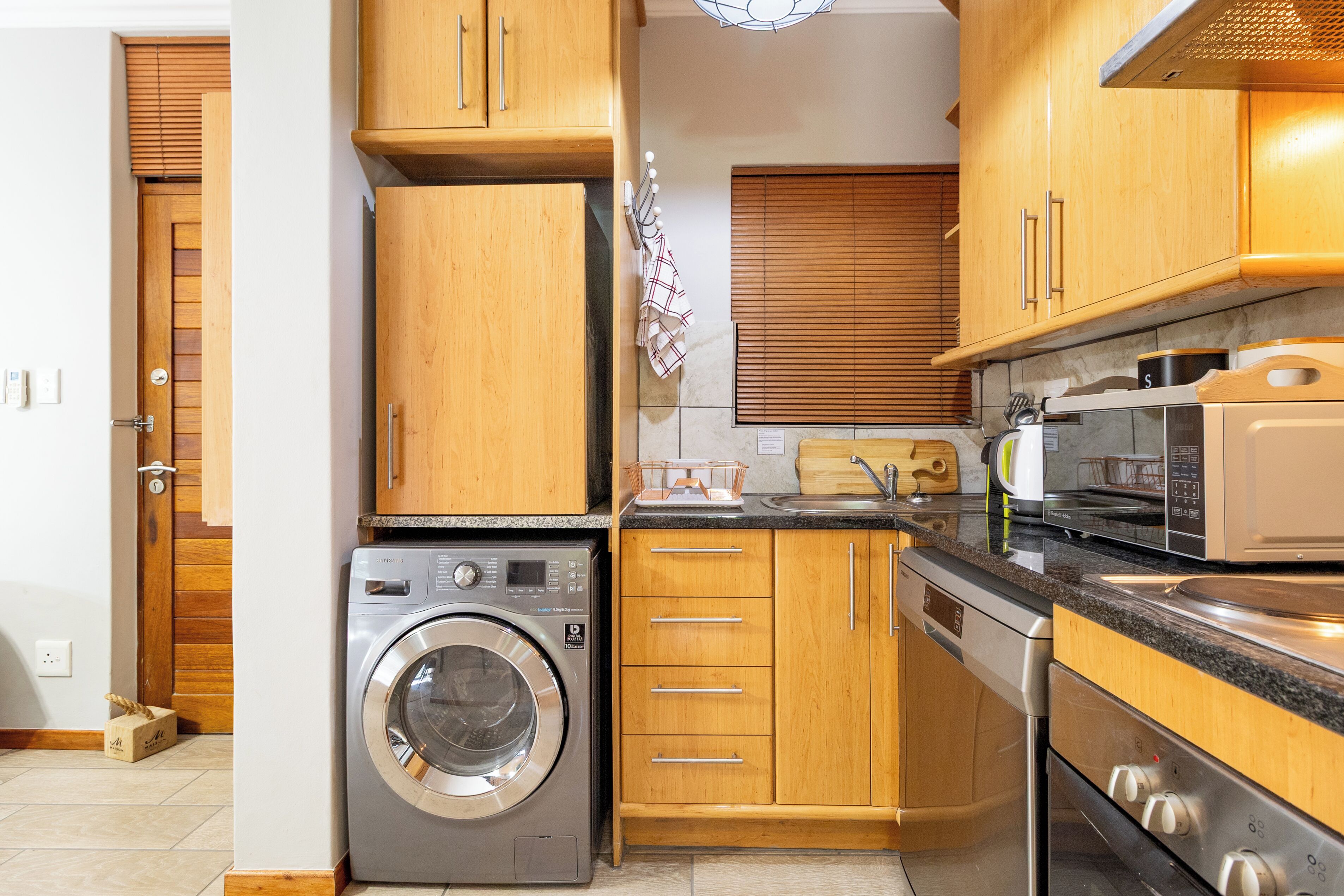 A compact kitchen with wooden cabinets, a washing machine, a dishwasher, and a microwave.