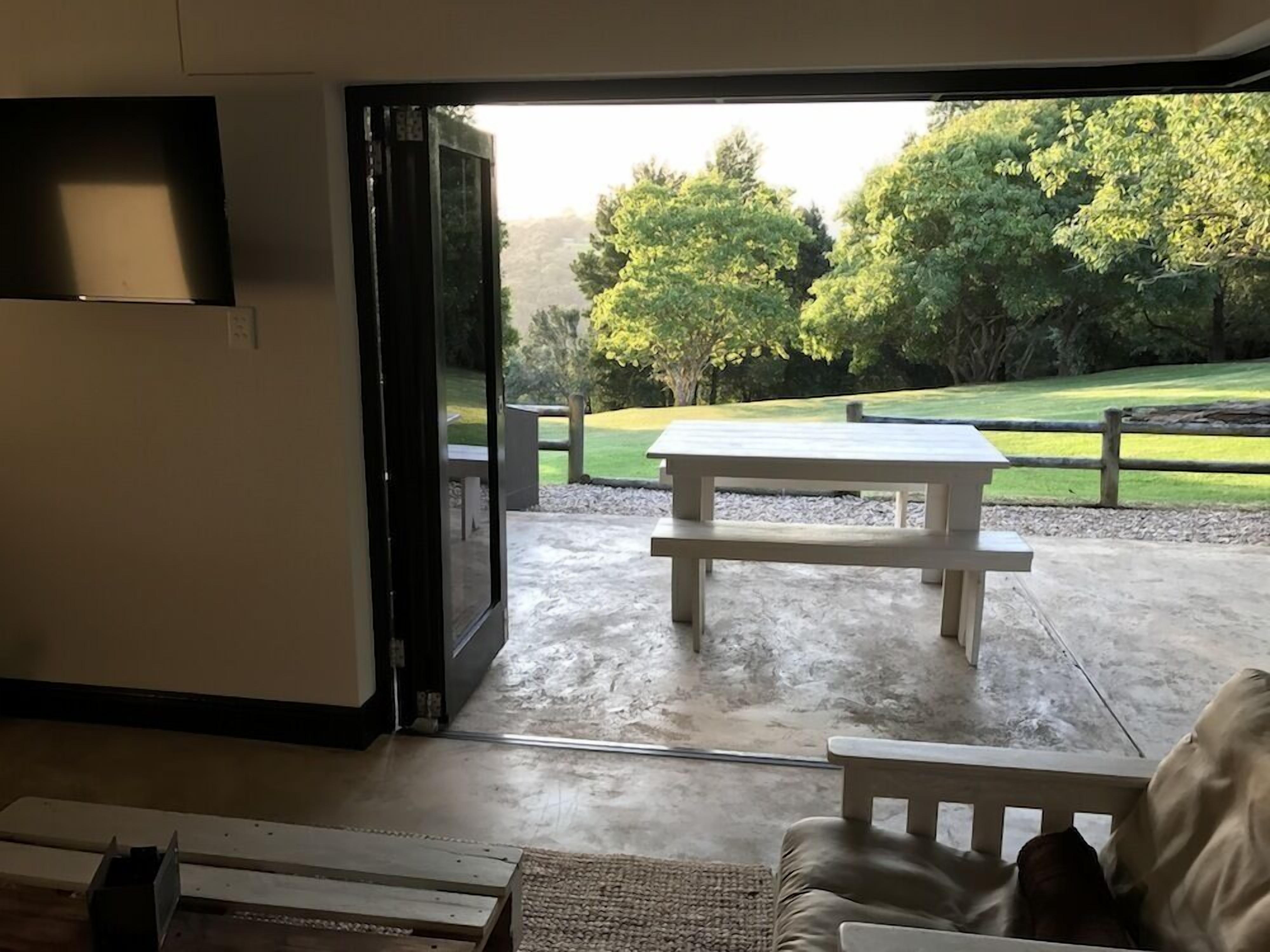 Une porte ouverte donne sur un patio équipé d’une table de pique-nique et offre une vue sur un espace verdoyant bordé d’arbres.