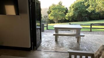 Une porte ouverte donne sur un patio équipé d’une table de pique-nique et offre une vue sur un espace verdoyant bordé d’arbres.