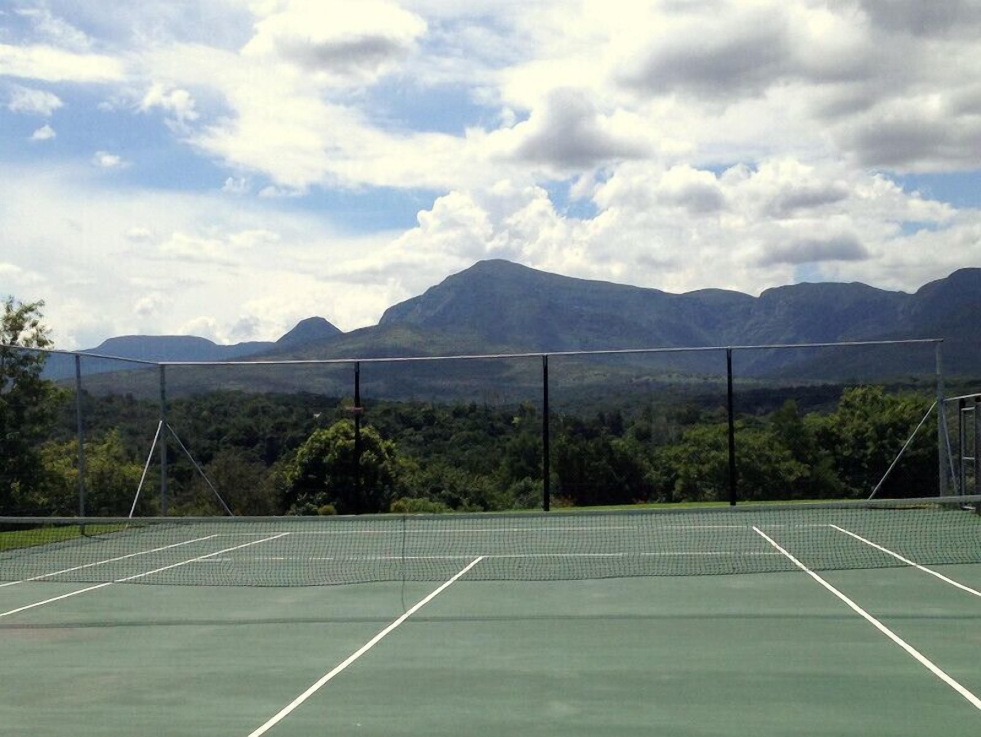 Una cancha de tenis con una red, rodeada por una valla metálica, y montañas al fondo.