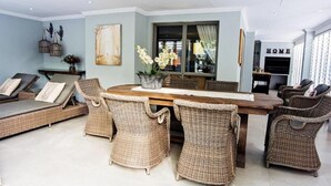 Een ruime woonruimte met een centrale houten tafel, omringd door rieten stoelen, een bank en een decoratieve vaas met bloemen.