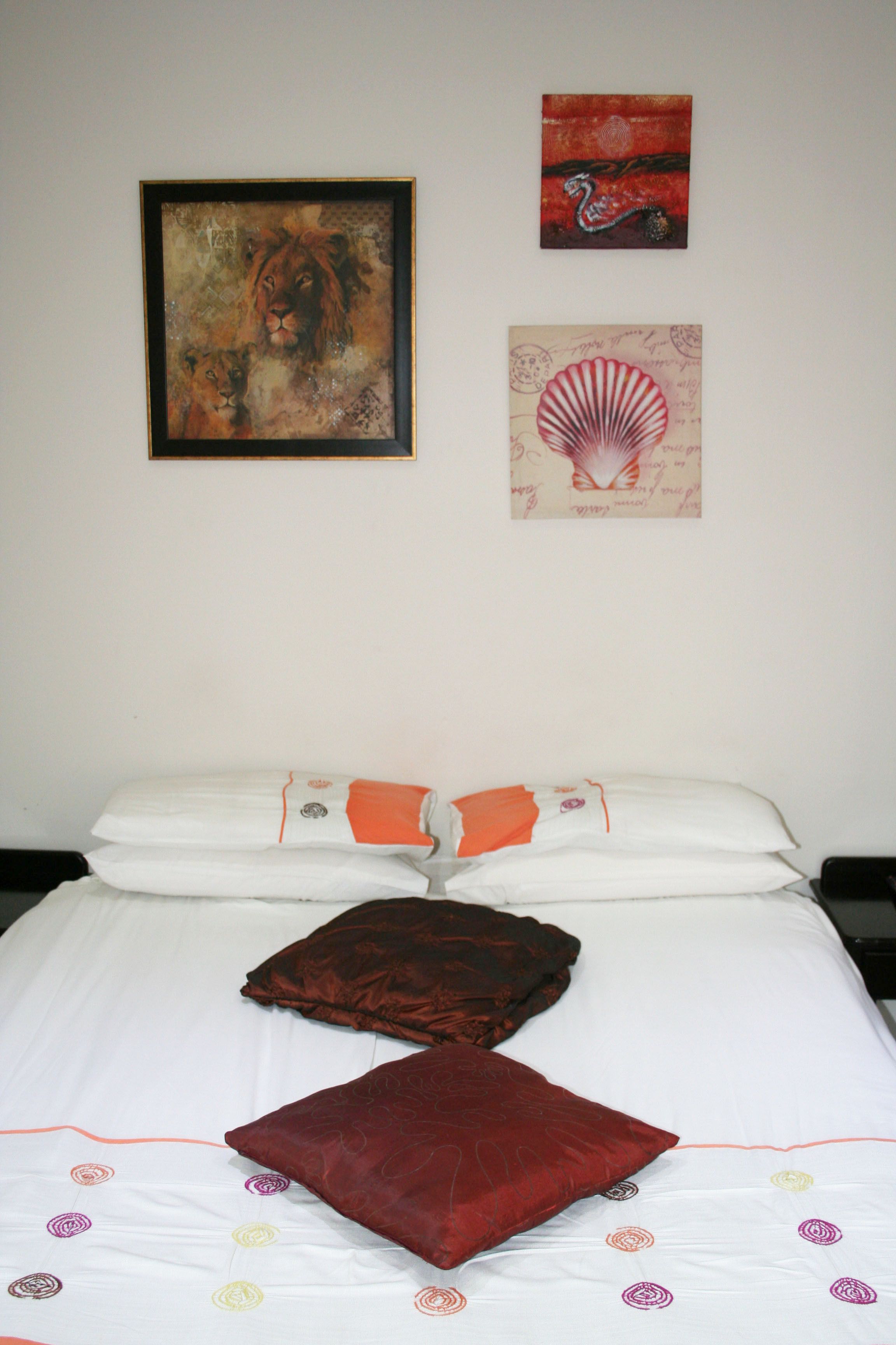 Een net opgemaakt bed met een wit-oranje dekbedovertrek, twee kussens en een rode sierkussen. Boven het bed hangen drie kunstwerken in lijsten.