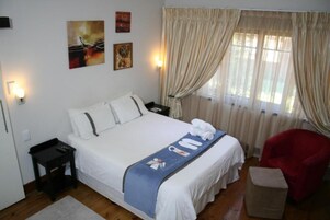 Una habitación de hotel con cama, mesita de noche, una silla roja y ventana con cortinas.