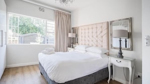 A bedroom with a large bed, a bedside table with a lamp, a window with curtains, and a chandelier. - Avemore Vredehof No 1- with backup power (Stellenbosch)