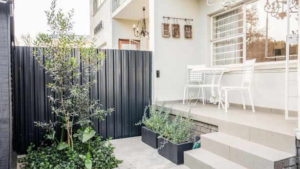 A modern outdoor patio with a black fence, white chairs, and a small garden area. - Avemore Vredehof No 1- with backup power (Stellenbosch)