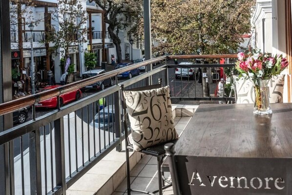 Un balcón con vista a una calle concurrida, una mesa con un florero y una silla con cojín estampado.
