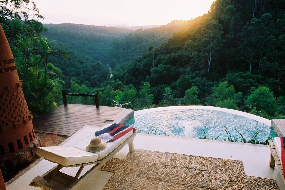 A poolside area with a lounge chair and a hat, surrounded by lush greenery and a view of a valley.