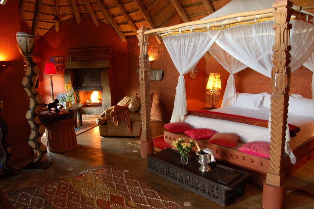 A four-poster bed with a canopy, a fireplace, a desk with a lamp, and a patterned rug.