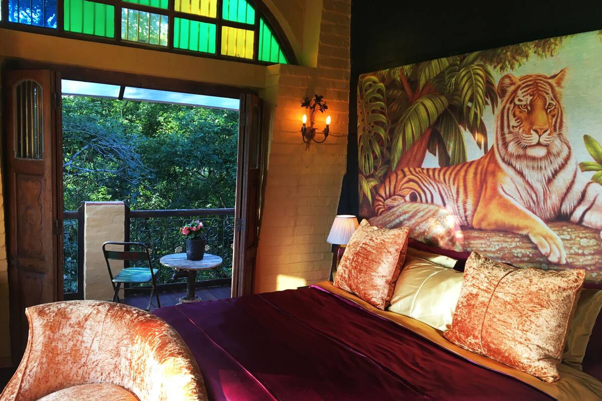A room with a bed, a view of greenery outside, a painting of a tiger, and a small table with a chair.