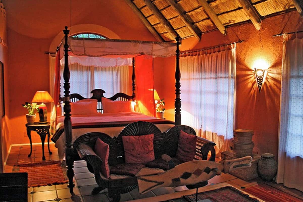 A four-poster bed with a canopy, a sofa with cushions, a lamp, and a wall-mounted light fixture in a room with a tiled floor and a thatched ceiling.