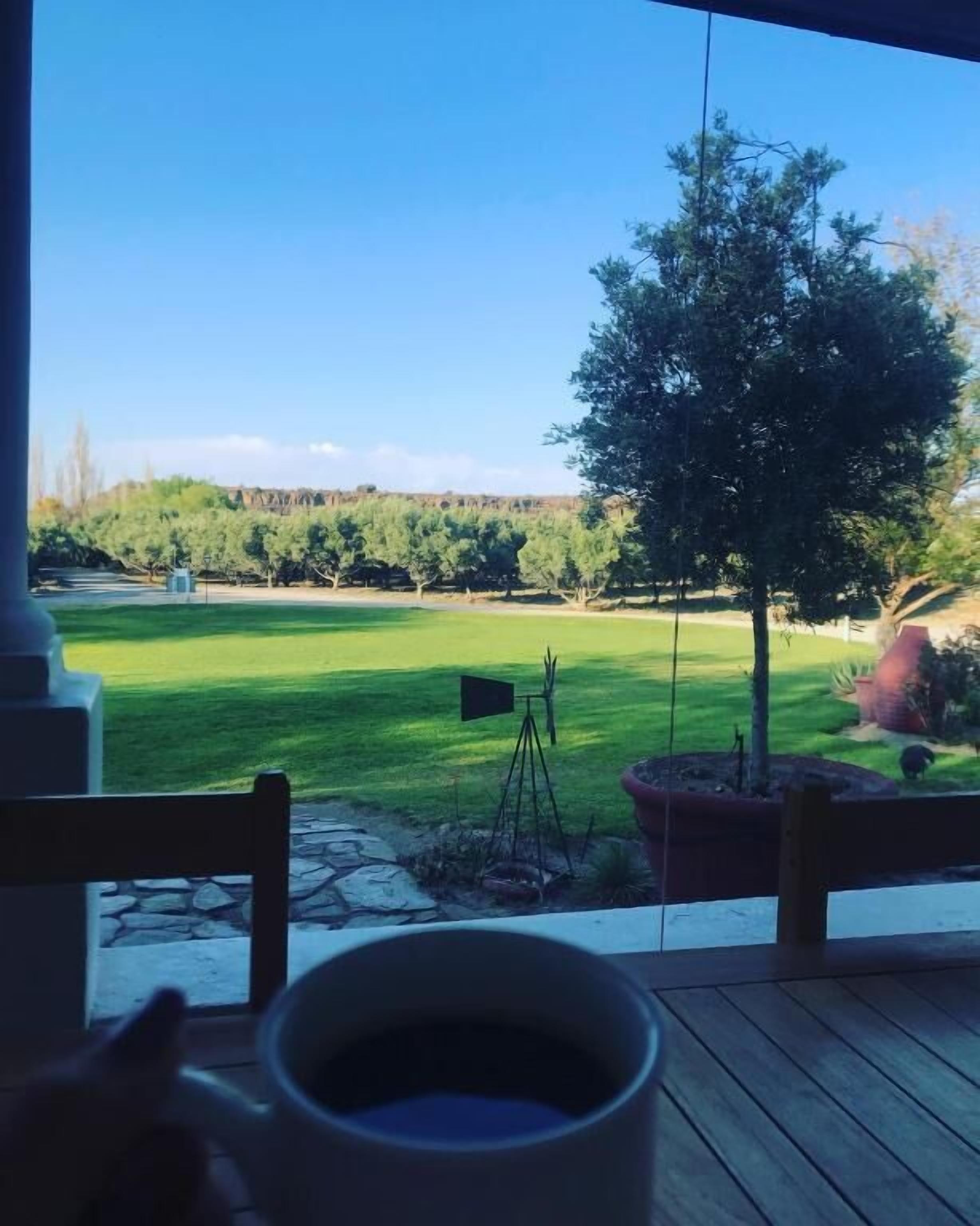 A view from a room with a wooden floor and a table with a cup, overlooking a green lawn with a flag, trees, and a distant hill.