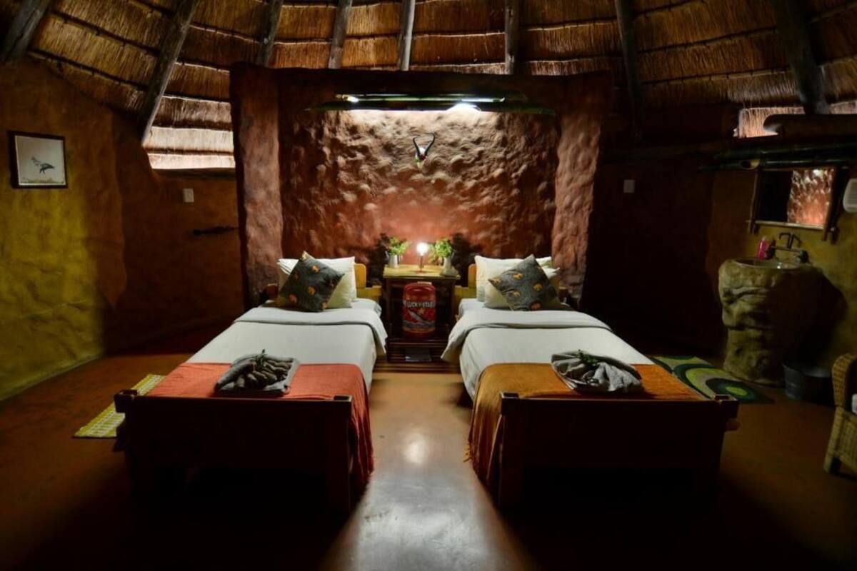 A room with two beds, a small table with a plant, and a thatched roof.
