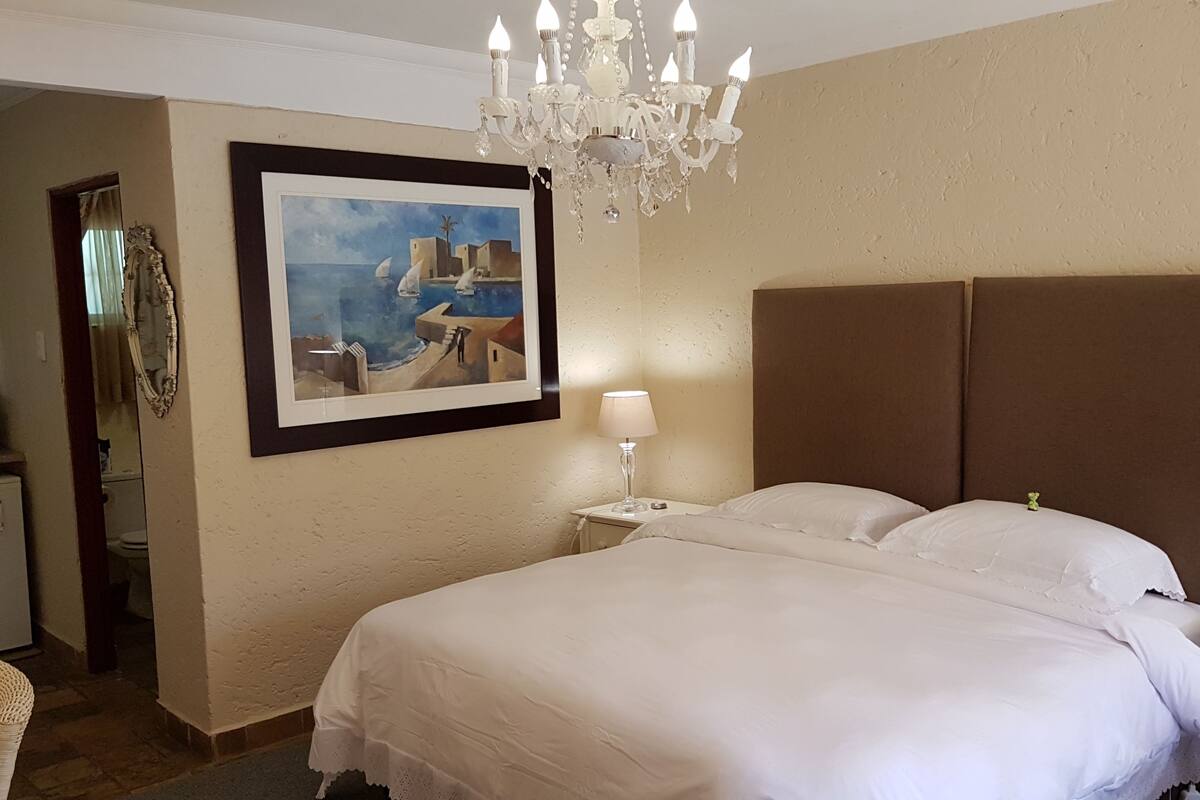 A hotel room with a large bed, a chandelier, a painting, and a small table with a lamp.