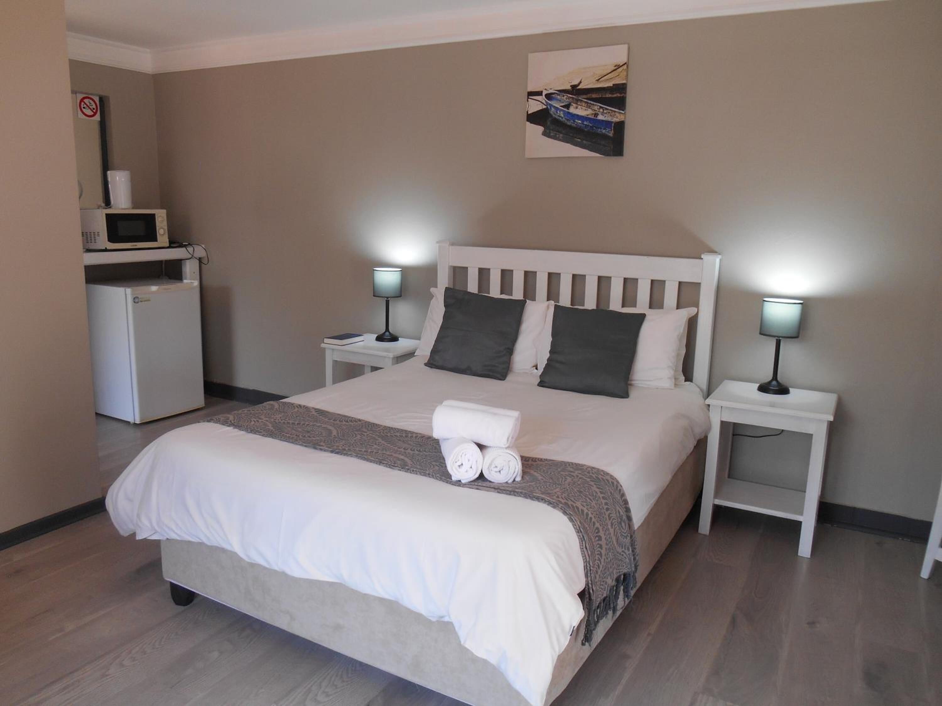 Cama bem arrumada com edredom branco, duas mesas de cabeceira com abajures, um frigorífico branco pequeno e uma moldura na parede.