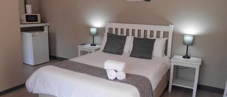 Cama bem arrumada com edredom branco, duas mesas de cabeceira com abajures, um frigorífico branco pequeno e uma moldura na parede.