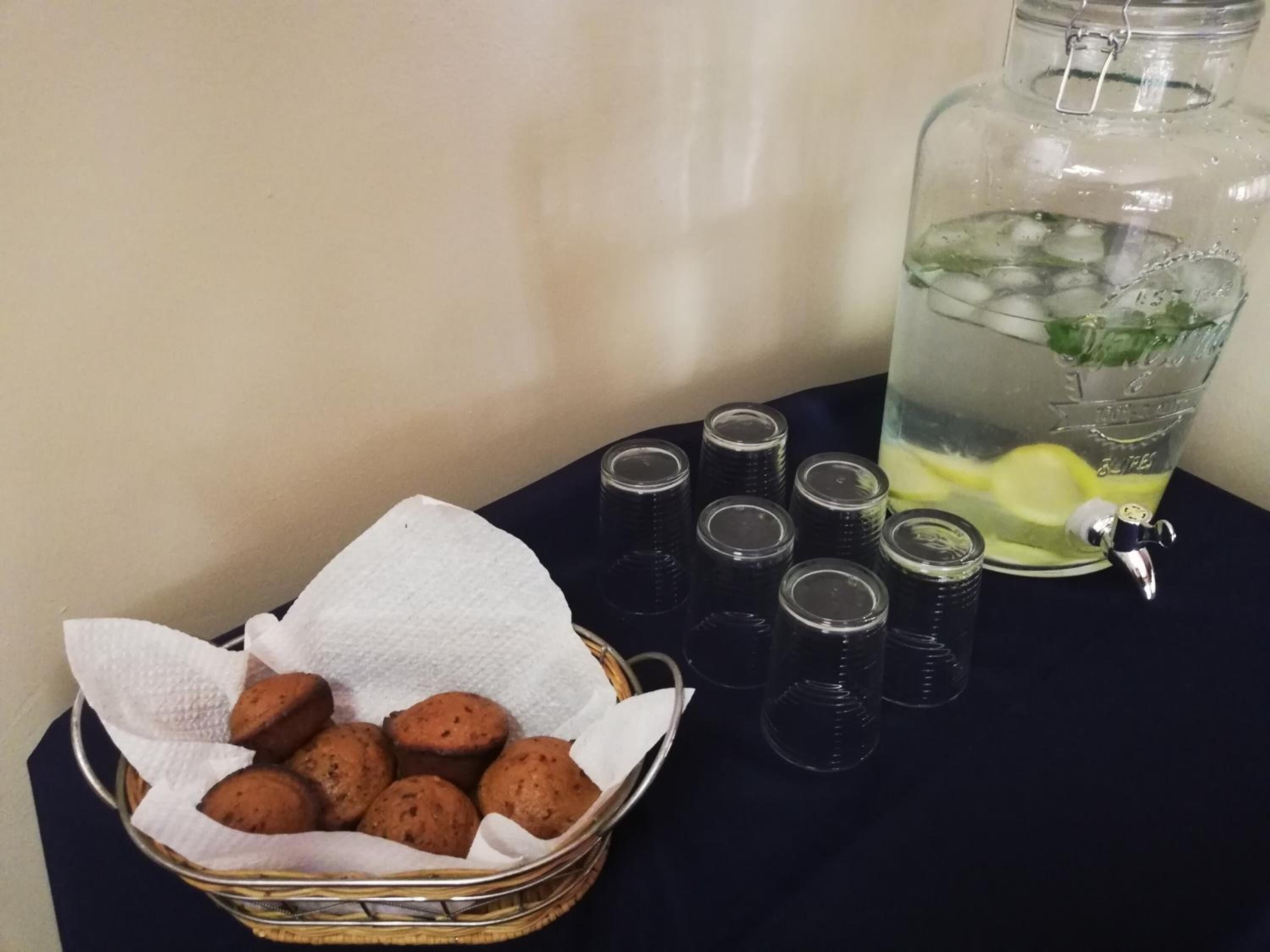 Una cesta de muffins, una jarra de vidrio con una rodaja de limón y varios vasos exprimidor sobre una mesa.