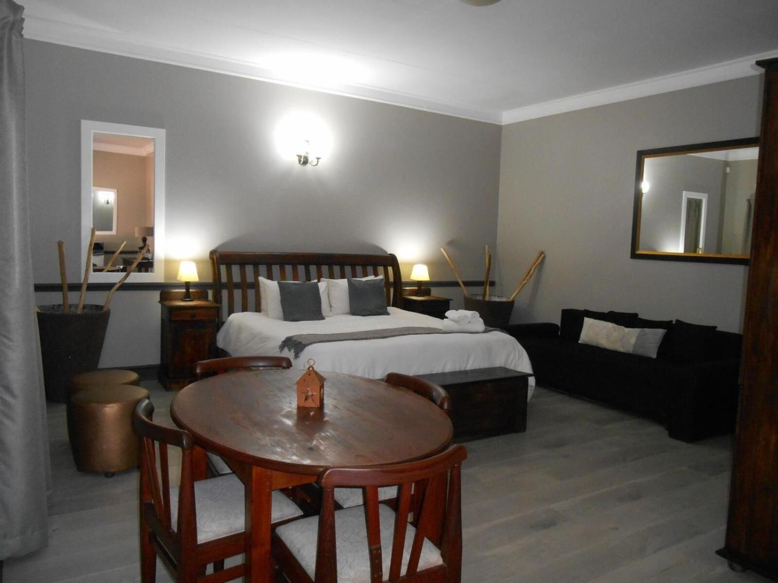 A hotel room with a bed, bedside tables, a wooden dining table with chairs, a sofa, and a mirror.