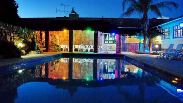 A pool area with a reflection of the building, a stone wall, and a palm tree. - Oasis Country Lodge (Matzikama)