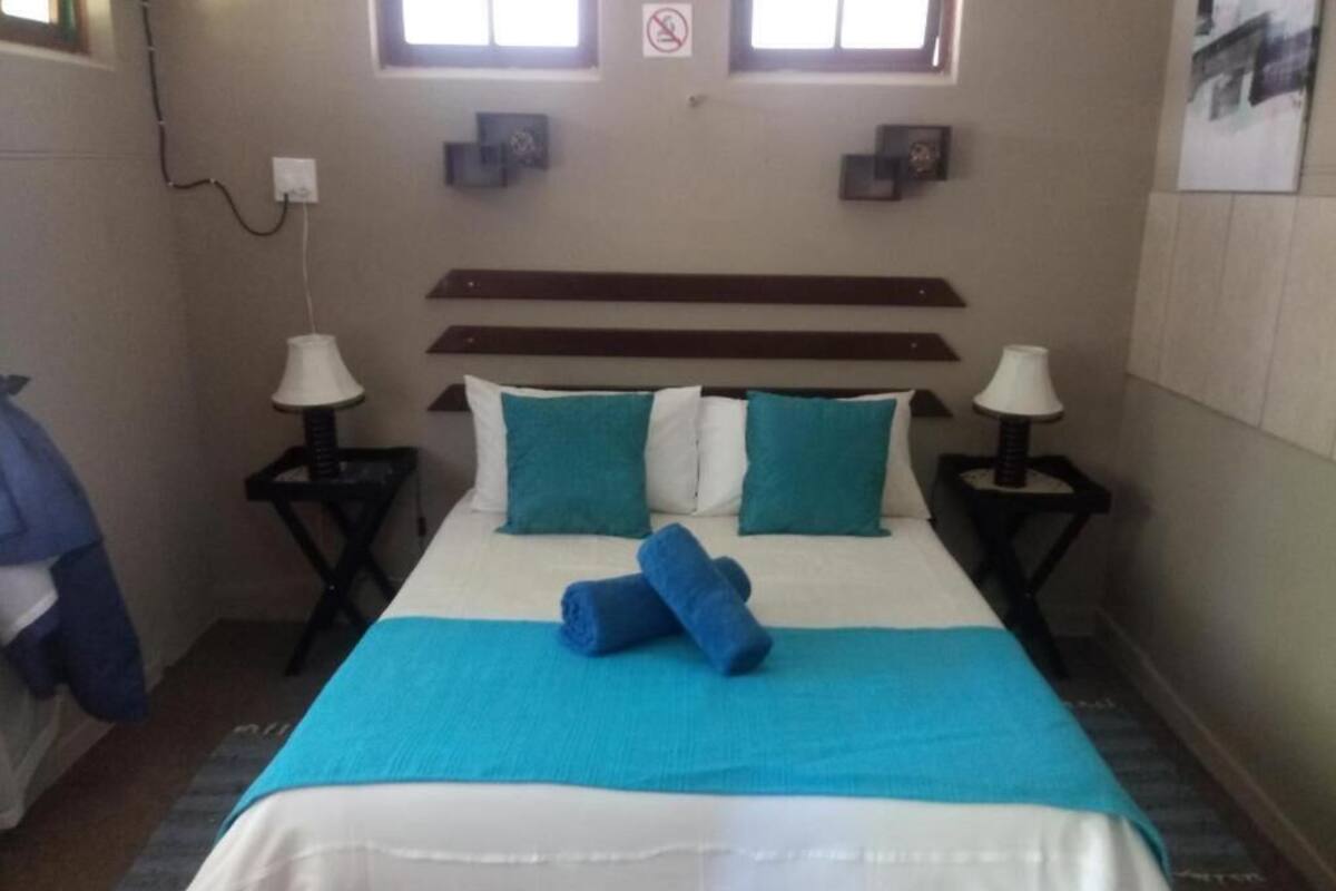 Una cama bien hecha con una colcha azul y blanca, dos almohadas y dos toallas dobladas. Hay dos mesitas de noche con lámparas, un estante en la pared sobre la cama y una ventana con vistas al exterior.