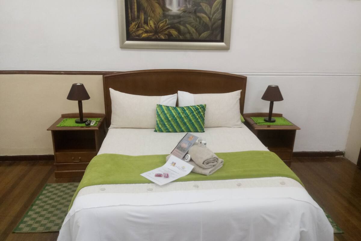 Una cama bien hecha con una manta verde, almohadas y una toalla doblada. Hay dos mesitas de noche con lámparas y un cuadro enmarcado sobre la cama.