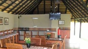A dining area with wooden tables and chairs, a television mounted on the wall, and a kitchen area in the background. - Leselo La Bontshi Lodge (Groblersdal)