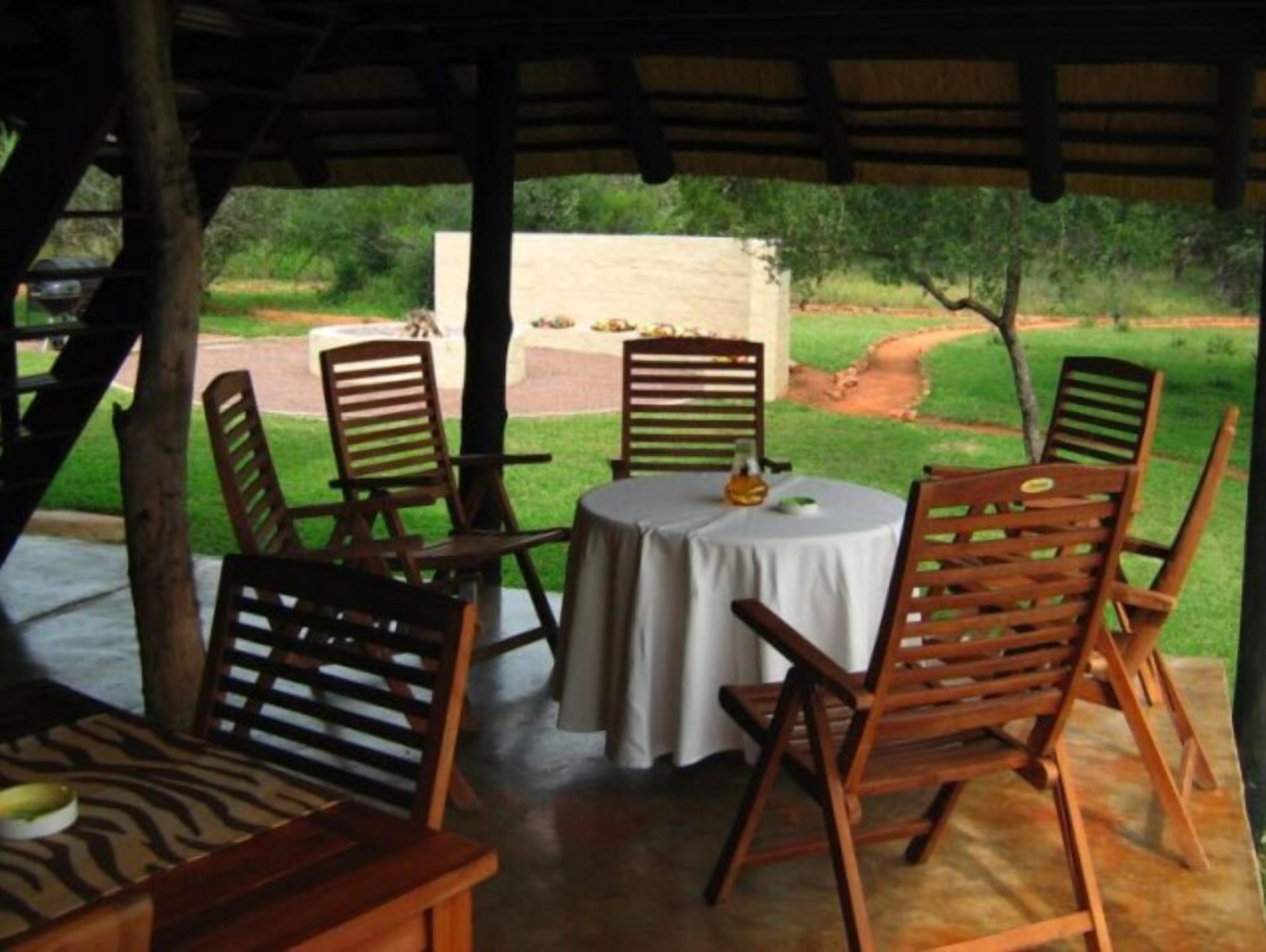 A covered outdoor seating area with wooden chairs and a table, overlooking a garden with a pathway and greenery.