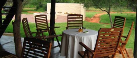 A covered outdoor seating area with wooden chairs and a table, overlooking a garden with a pathway and greenery.