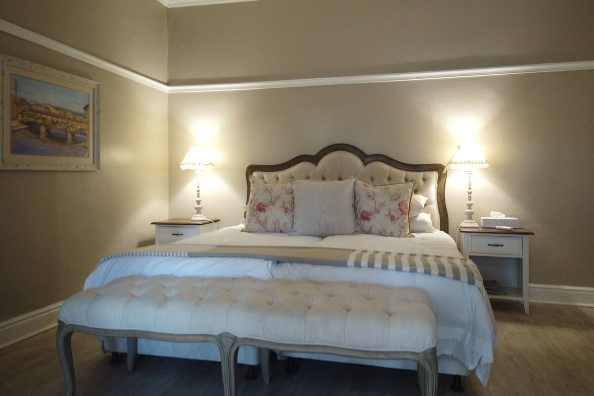 A bedroom with a large bed, two bedside tables with lamps, a bench at the foot of the bed, and a painting on the wall.