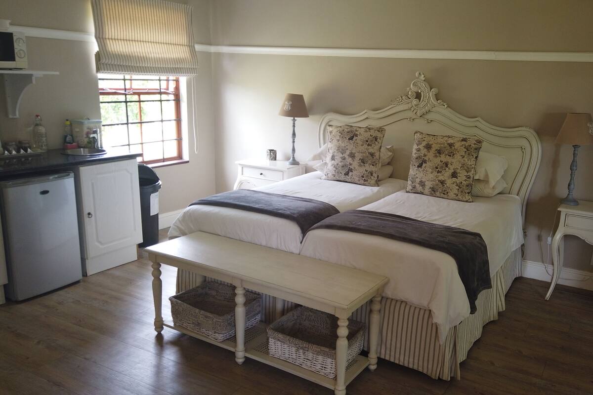 A bedroom with a double bed, bedside tables, a bench, a small refrigerator, and a window with blinds.