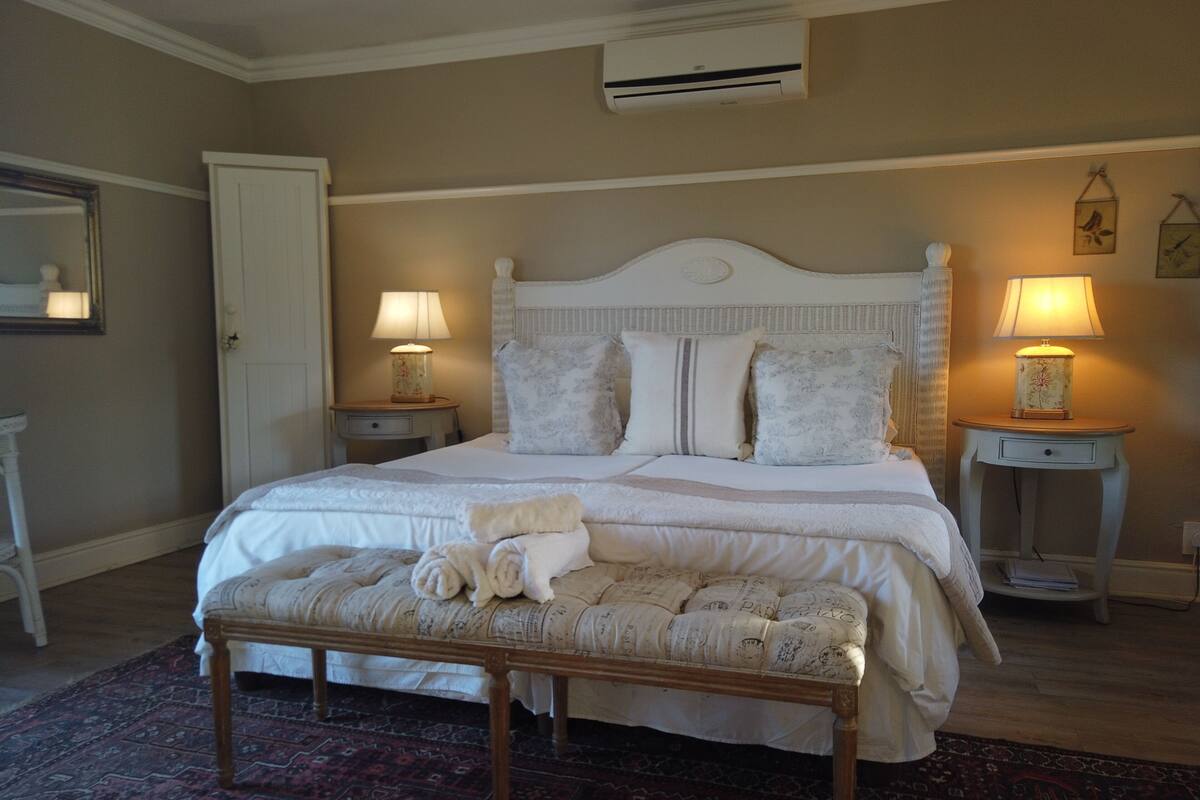A neatly arranged bedroom with a bed, two bedside tables with lamps, a bench with towels, and an air conditioning unit on the wall.