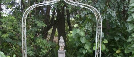 A metal garden bench with a decorative top arch, surrounded by lush greenery and a statue.