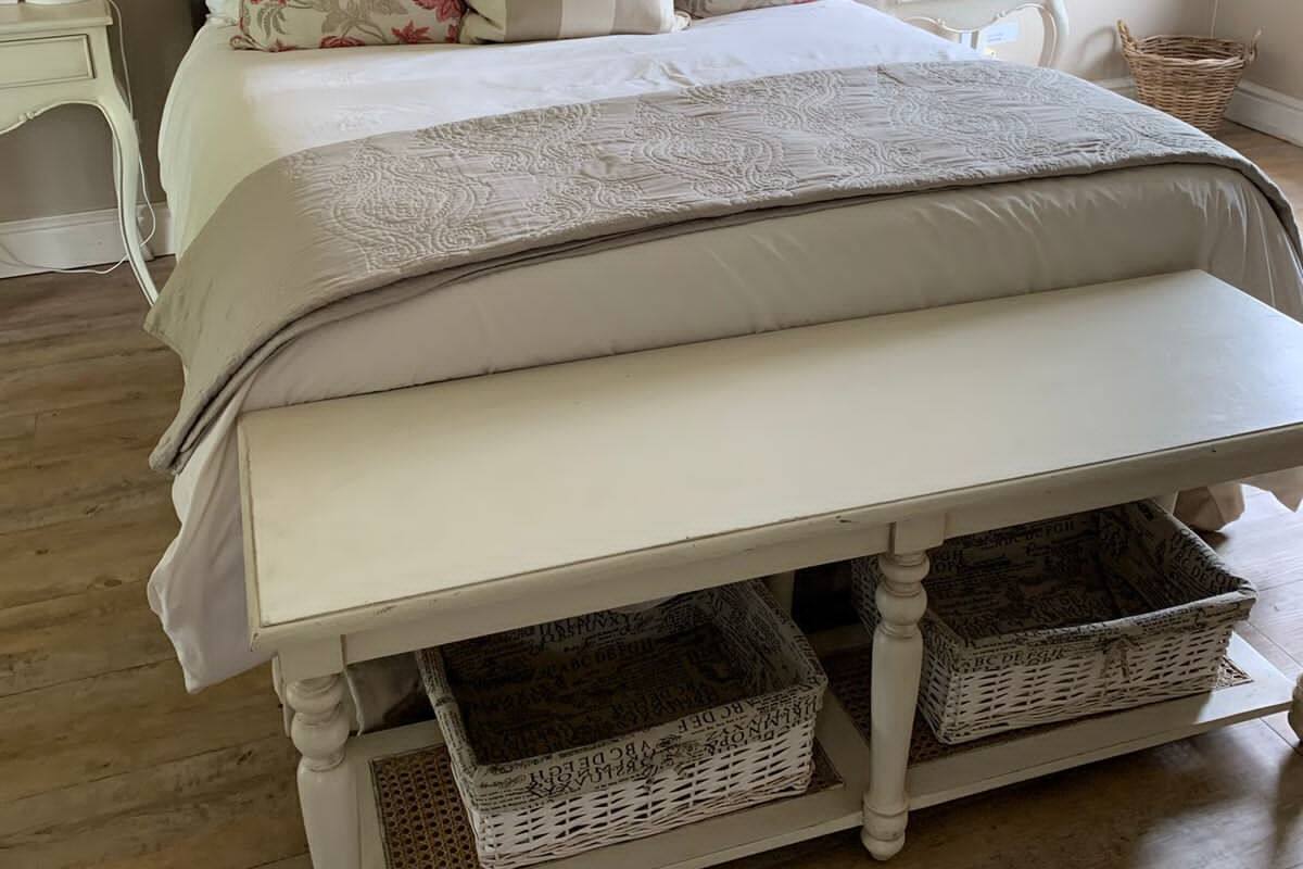 A bedroom with a tufted headboard, a large bed with floral and striped pillows, a bedside table with a lamp, and a wicker basket under the bed.