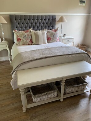A bedroom with a tufted headboard, a large bed with floral and striped pillows, a bedside table with a lamp, and a wicker basket under the bed. - Happy Lands Self Catering B&B (Addo)