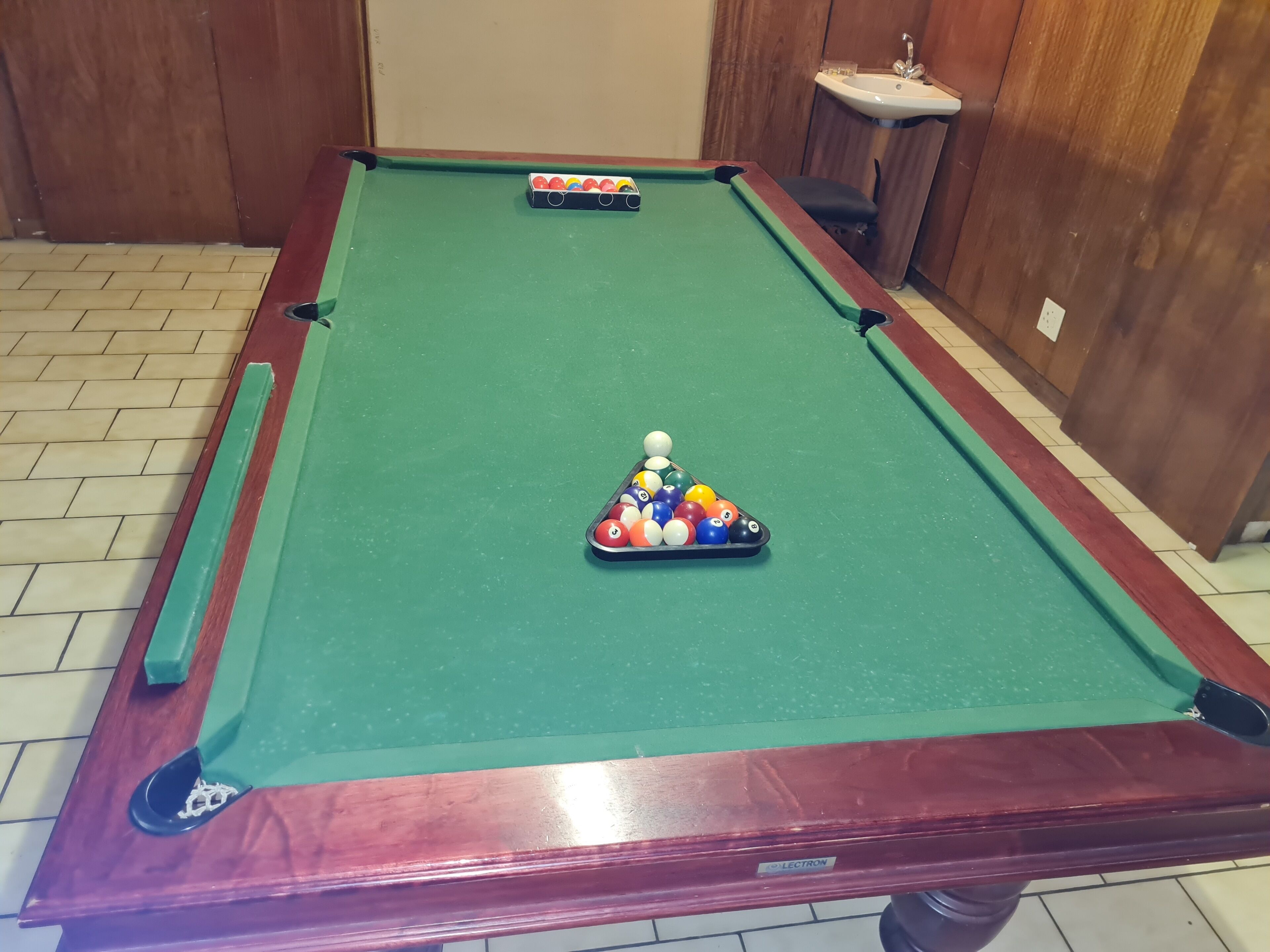 A pool table with a triangular rack of billiard balls in the center, a pool cue, and a small sink in the background.