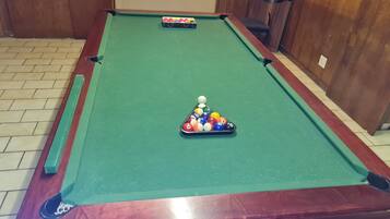 A pool table with a triangular rack of billiard balls in the center, a pool cue, and a small sink in the background.