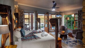 A spacious room with a large bed, a wooden side table, a small round table with a bowl of fruit, and a view of the outdoors through large windows. - Dungbeetle River Lodge (Colchester)