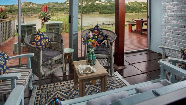A deck with wicker furniture, a wooden table, and a view of a lake and hills. - Dungbeetle River Lodge (Colchester)