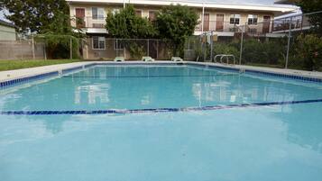 Una piscina rectangular con baldosas azules, rodeada por una valla de metal. Se observan árboles y un edificio con varias ventanas y puertas al fondo.